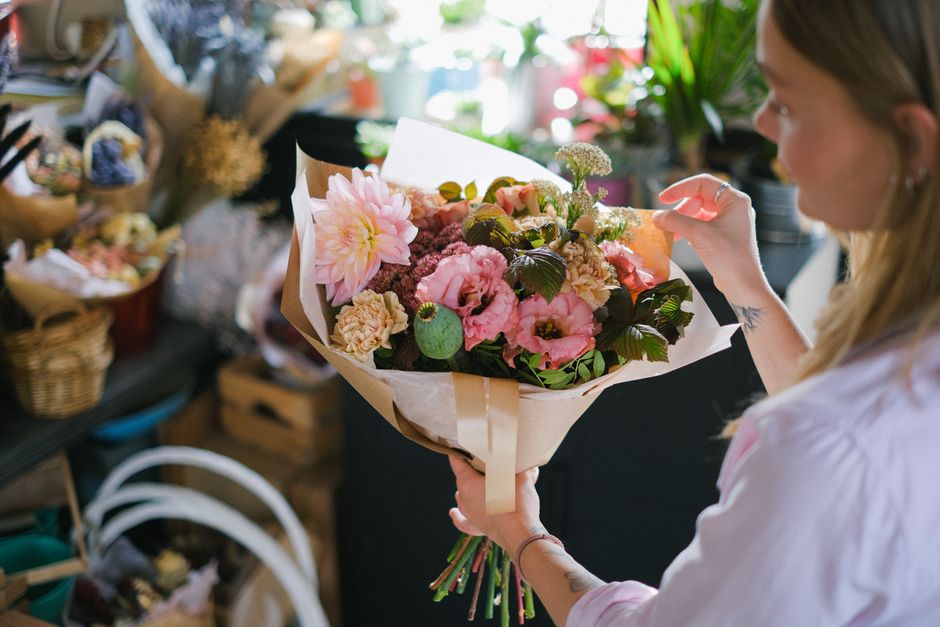 A woman with long blonde hair, wearing a white shirt, is holding a large bouquet of fresh flowers wrapped in beige paper with a satin ribbon. The bouquet includes pink dahlias, pink lisianthus, beige