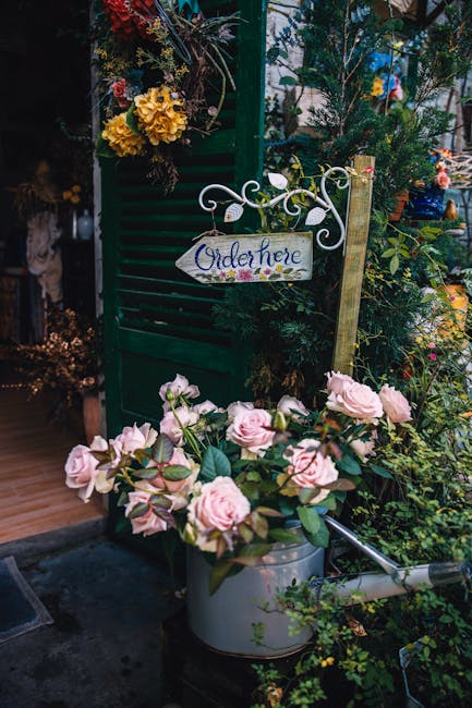 A display of fresh light pink roses with lush green foliage arranged in a metal watering can, situated near a vibrant green wooden lattice decorated with yellow and dark purple flowers. The scene is s