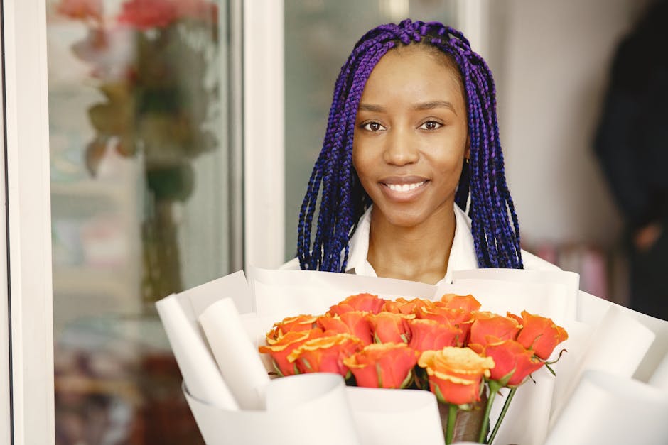 A smiling woman with long, purple braided hair is holding a vibrant bouquet of fresh roses that are orange with pinkish-yellow edges, wrapped in white floral paper. She is standing inside, possibly at