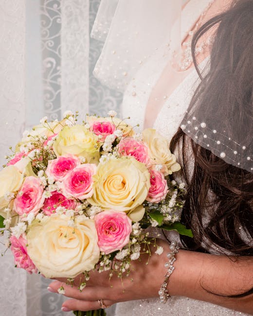 A bride holds a floral bouquet featuring large, creamy white roses and smaller pink roses, complemented by delicate white baby's breath and greenery. The bouquet is arranged in a rounded, lush style a