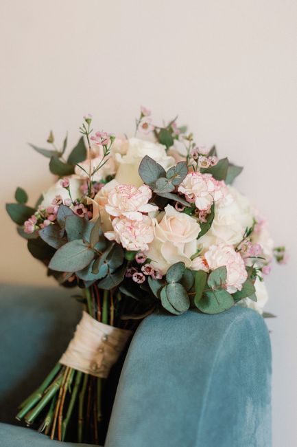 A floral bouquet resting on the armrest of a teal upholstered chair, featuring a harmonious arrangement of soft pink roses, pale pink carnations, and small pink spray roses interspersed with eucalyptu