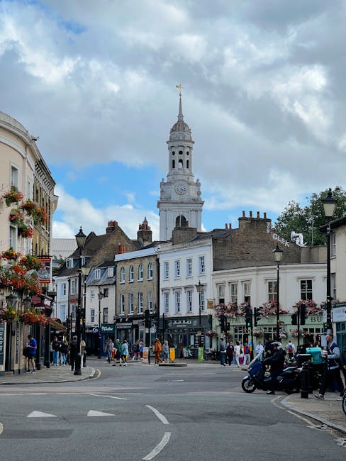 A bustling street scene in Barnsbury N1 featuring a variety of pedestrians and cyclists. In the background, a prominent historic clock tower with a white facade, a gold weather vane, and a spire rises