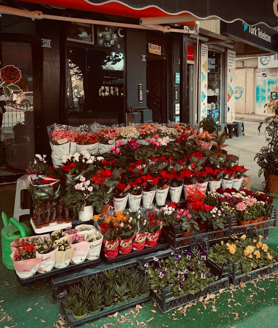 An outdoor display of fresh flowers arranged in white, red, pink, and black containers and buckets on a black metal table in front of a florist shop. The arrangement includes a variety of flowers such