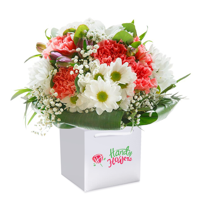 A lavish bouquet, meticulously arranged and upright in a stark white gift bag, radiates with colour and artistry in the light of Barnsbury. The dominant coral-pink and fiery red carnations, their ruffled petals layered in soft profusion, set the scene for an uplifting display. Intertwined among the carnations are clean white daisies with striking lime-green centers that bring an immediate touch of cheer-echoing the brightness of Barnsbury Park on a sunny day. Sprigs of ethereal white baby's breath introduce cloud-like softness, weaving throughout the arrangement with feathery grace. Bursts of deep green foliage, their glossy leaves forming a graceful arc around the base, provide contrast and assurance, offering a strong, natural foundation to the fullness of the bouquet. Subtly located on one side, several tightly closed purple-toned buds add visual intrigue and the promise of unfurling beauty. The bouquet's balanced, rounded silhouette is displayed in a gift bag designed for easy giving-its white ribbon handles adding both form and function. The whole image is set against a perfect white background, emphasising the bouquet's freshness and the skilled composition that Florist Barnsbury brings to each delivery, whether for birthdays, anniversaries, or simply to surprise a loved one near Canonbury Square. Every stem and detail reflects the lively, generous spirit that defines Barnsbury's floral artistry.